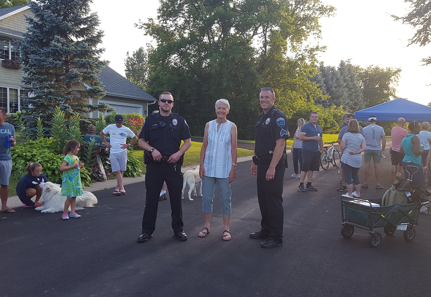 Shannon between two police officers—pose for a photo at a neighborhood community event with attendees and dogs nearby.