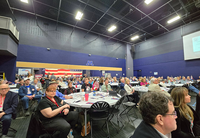 Large gym-style event hall filled with round tables and attendees, many wearing badges, patriotic decor, and American flags on display.