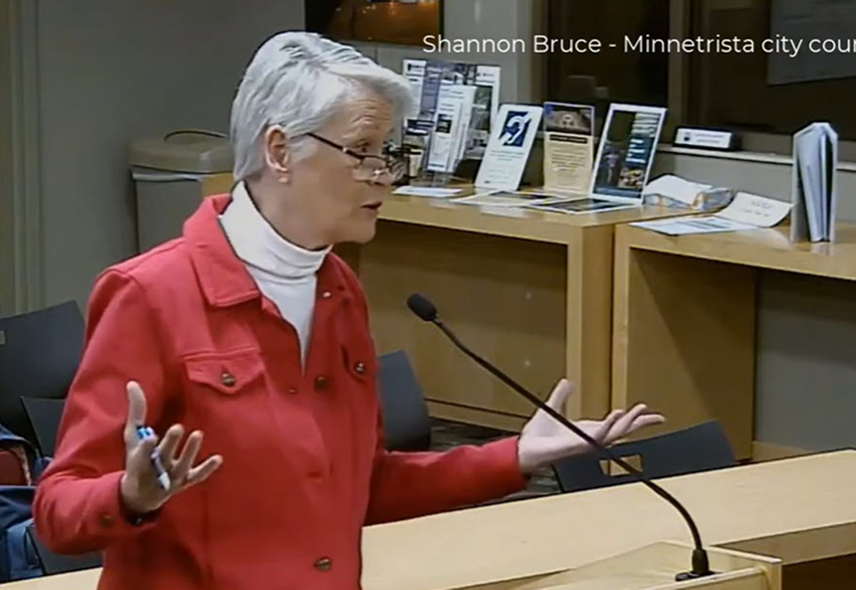 Shannon Bruce speaks at a podium with a microphone in a city council chamber, gesturing with her hands.