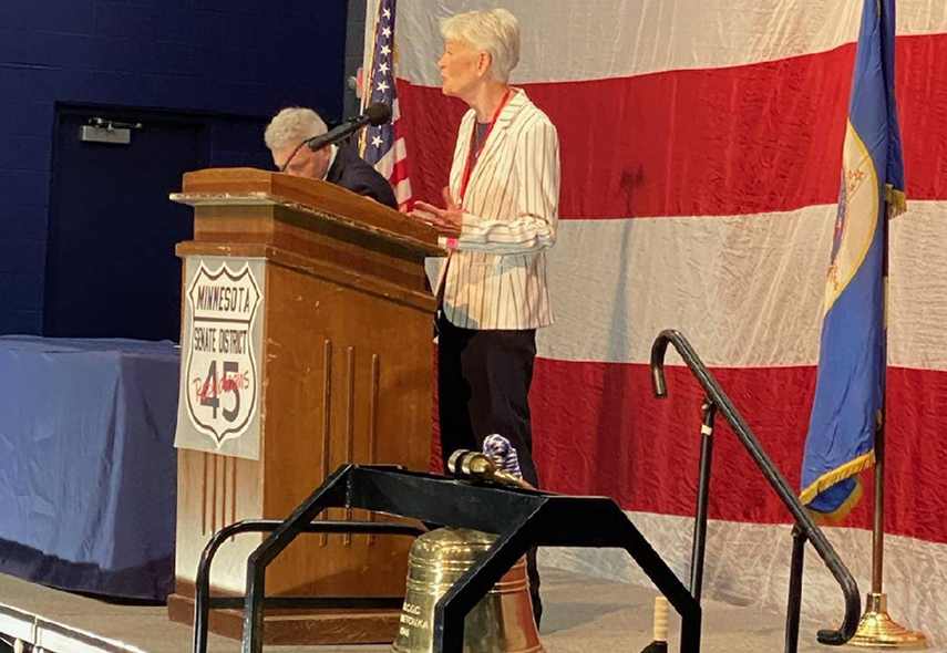 Shannon Bruce speaks at a wooden podium in front of an American flag and a Minnesota flag.