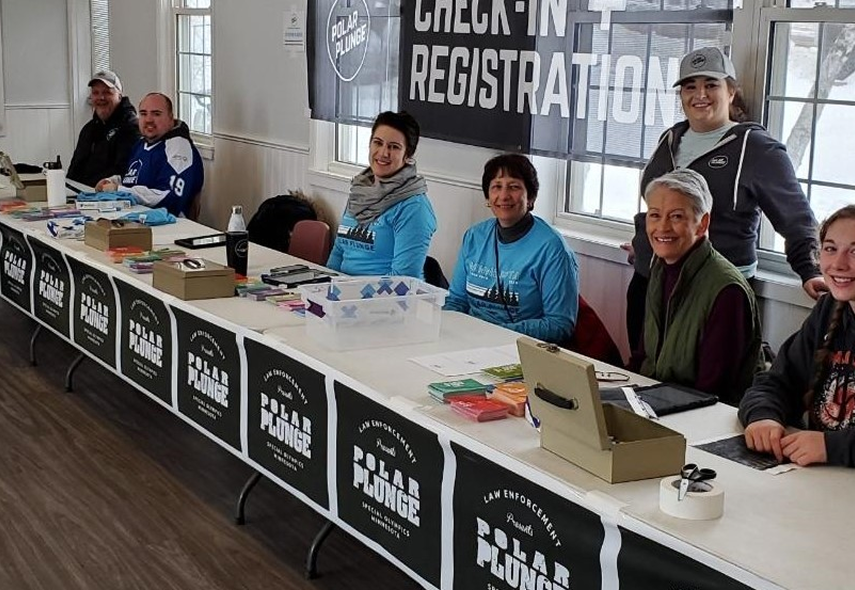 Volunteers sit at a long registration table under a 'Check-In Registration' banner for a Polar Plunge event, with boxes and papers in front of them.